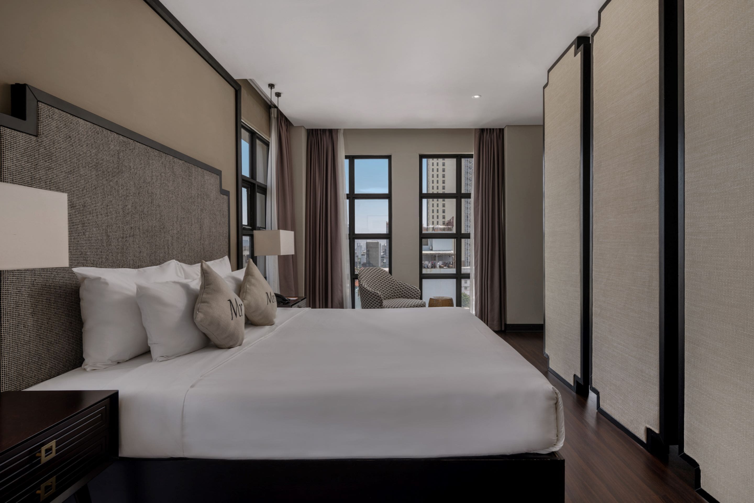 A modern hotel room with a large bed, a nightstand, a lamp, and a view of the city through large windows.