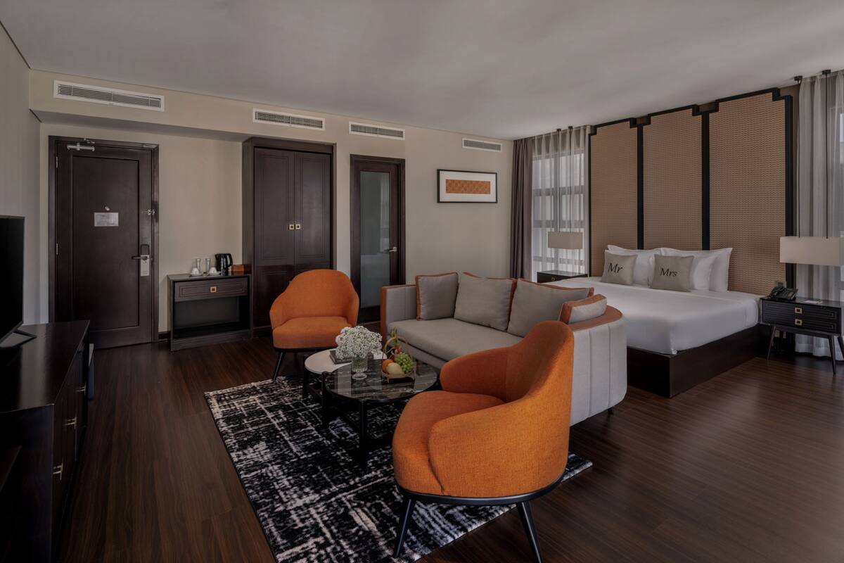 A hotel room with a bed, two orange chairs, a sofa, a coffee table, a TV, and a desk.