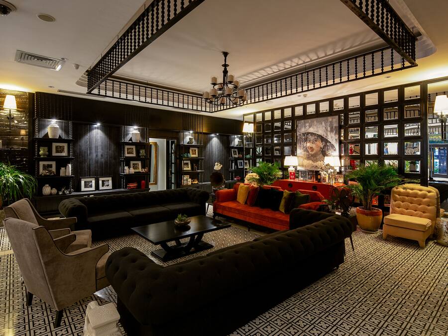 Un salón amplio con un sofá negro, un sofá rojo y un sillón amarillo. Se observan varias estanterías llenas de libros y objetos decorativos. En la pared se ve un cuadro de gran tamaño y, al fondo, una escalera con barandilla.