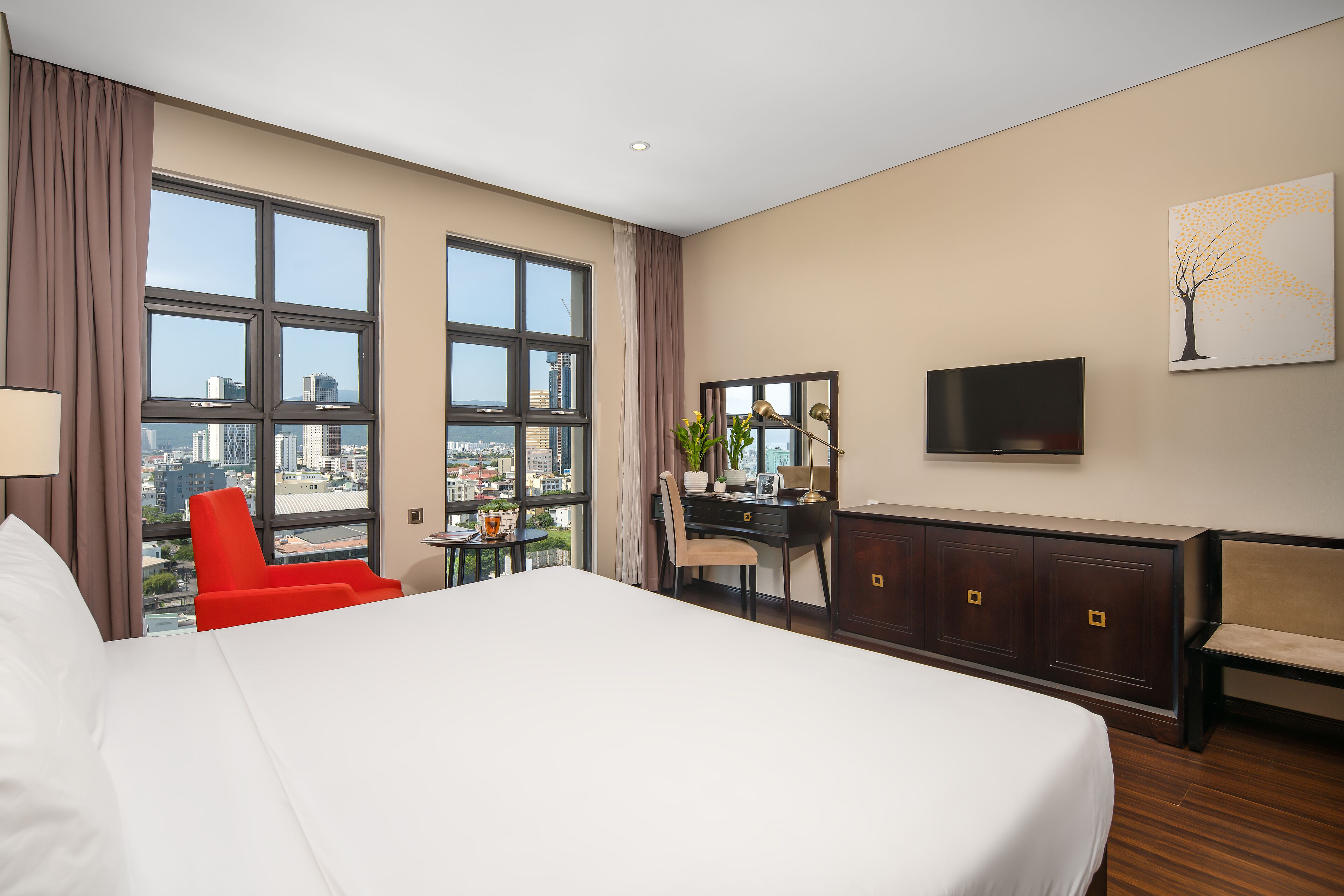 Habitación de hotel con una cama grande, un escritorio con silla, televisión y vistas a la ciudad a través de amplios ventanales.