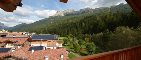 Vista desde un balcón de madera con vistas a un pueblo con casas tradicionales, un parque infantil y una cadena montañosa.