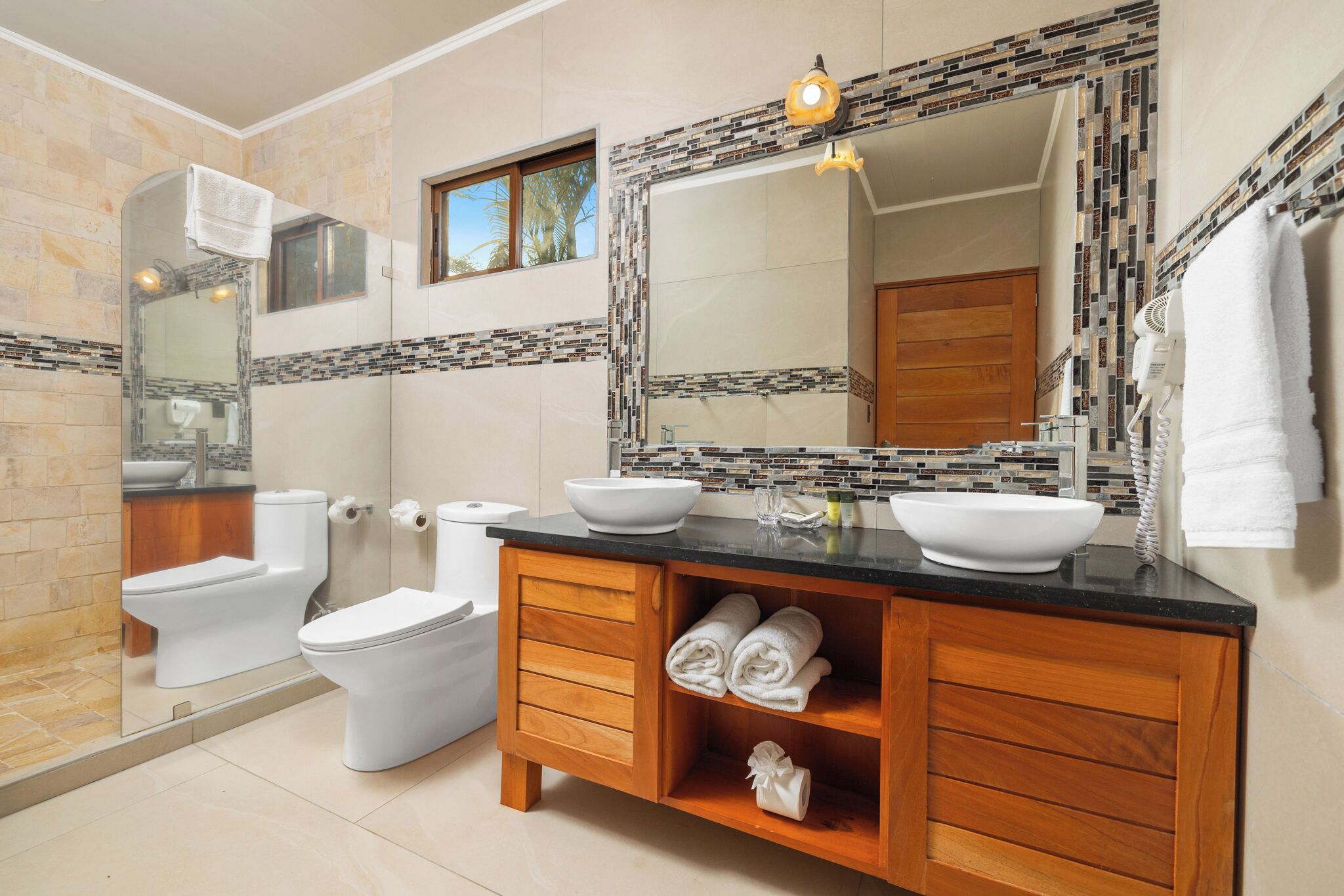 A bathroom with a large mirror, two sinks, a toilet, and a shower area.