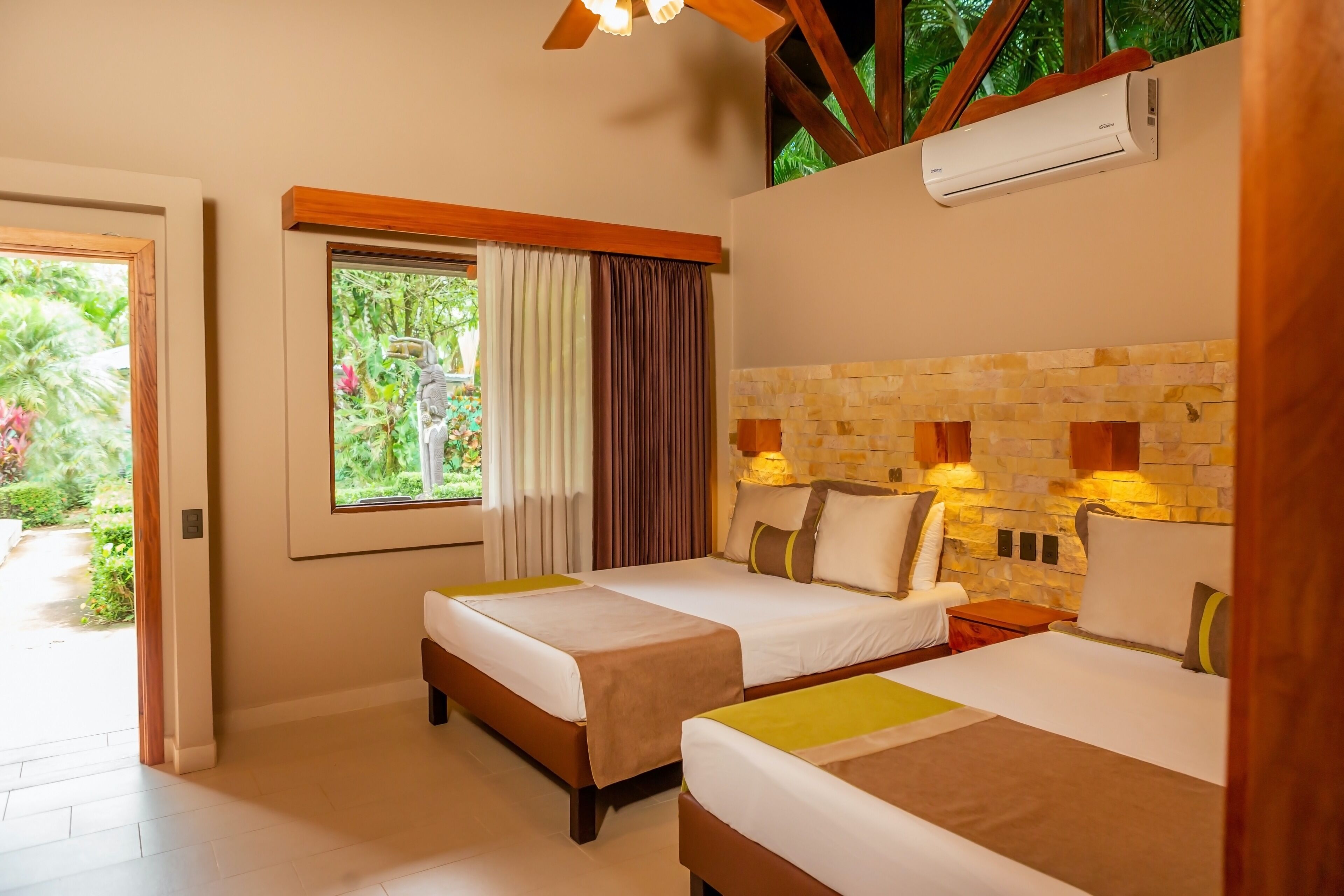 Habitación de hotel con dos camas, una pared de piedra, un equipo de aire acondicionado y una ventana con cortinas.