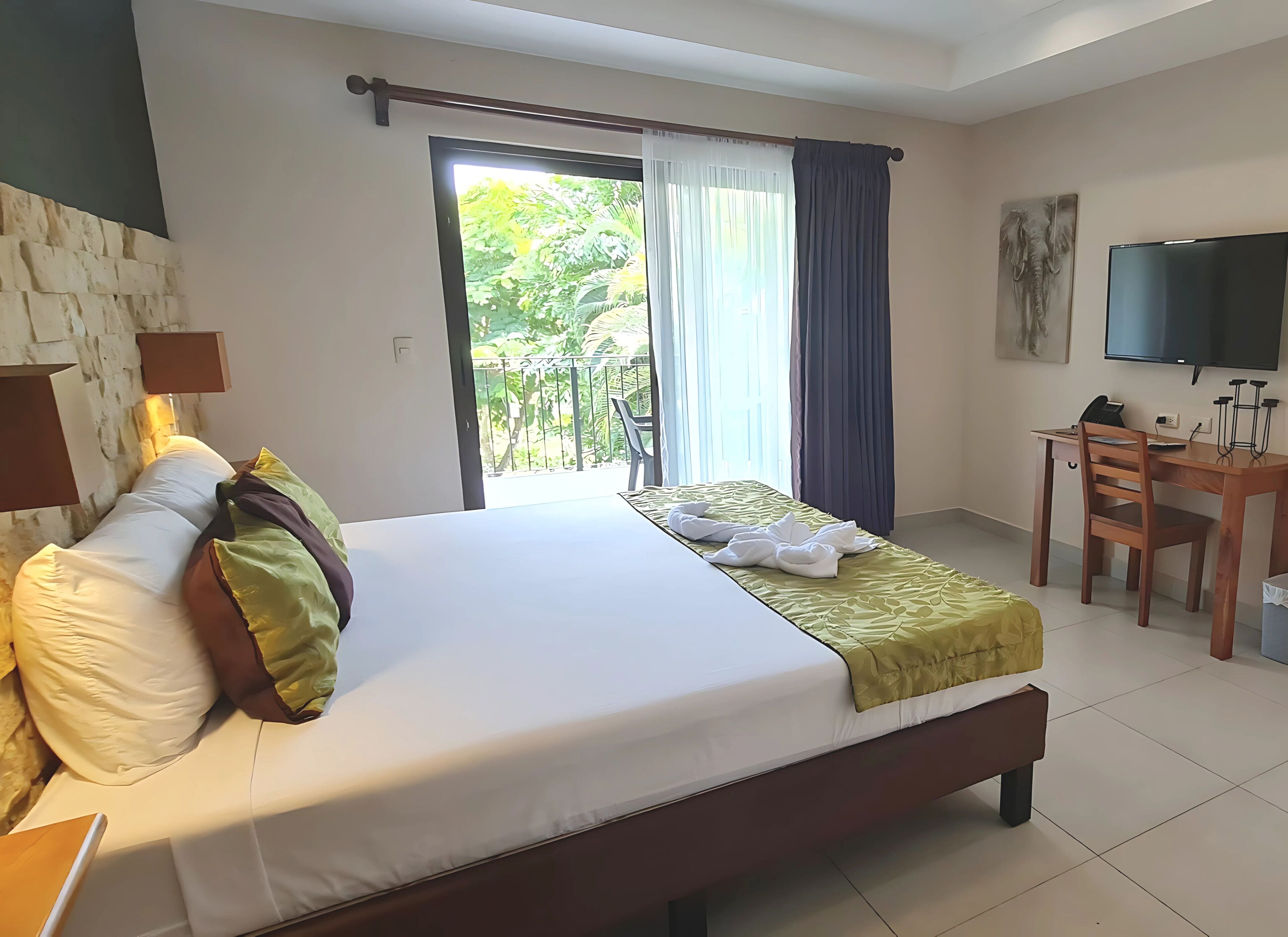 Habitación de hotel con cama, escritorio con silla, televisor y balcón con cortinas.