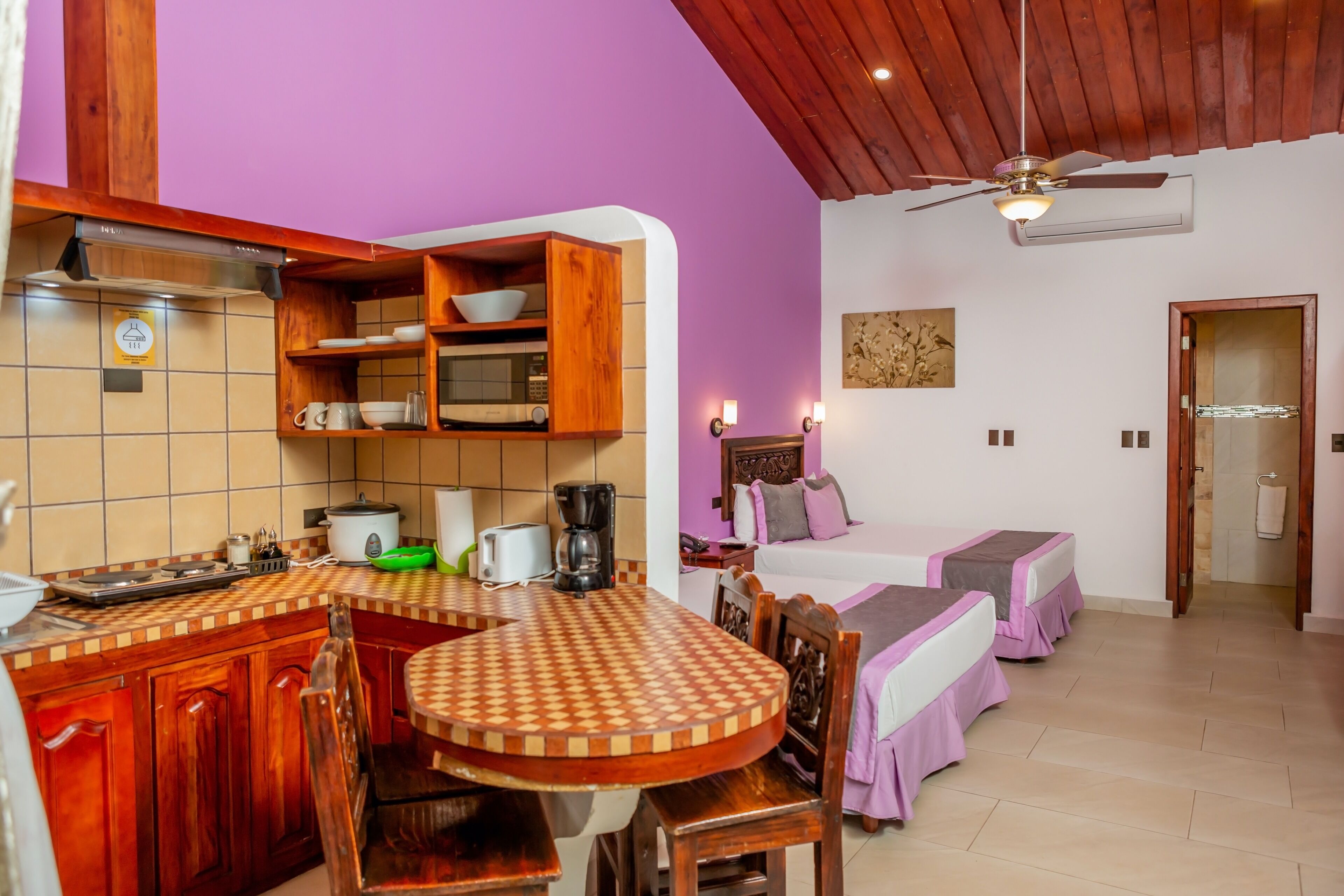 A kitchen with wooden cabinets, a microwave, and a coffee maker. There is a dining table with chairs and a bed with a purple bedspread.