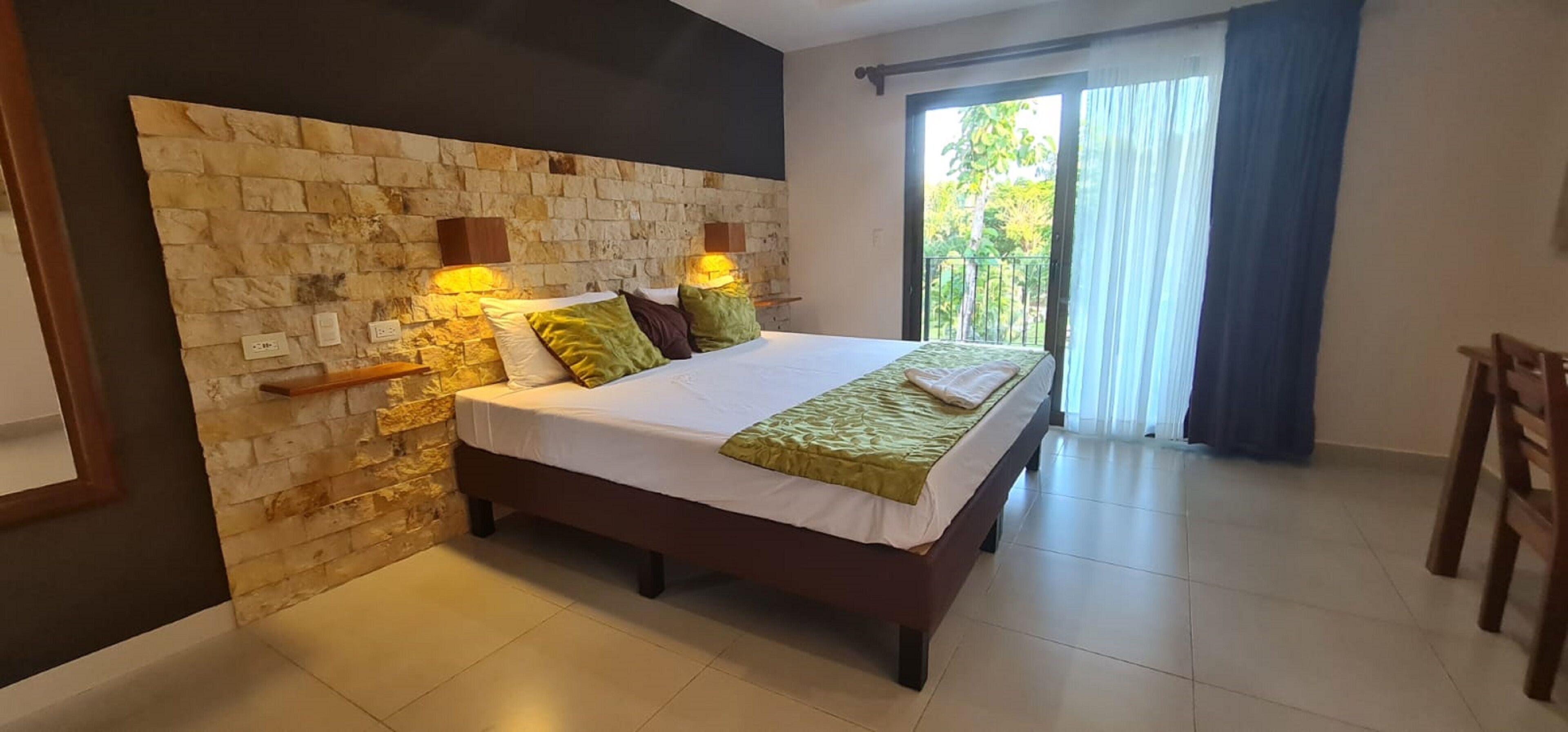 A bedroom with a stone headboard, a bed with green and white bedding, and a balcony with a view of greenery.