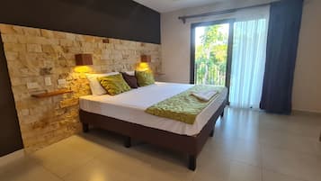 A bedroom with a stone headboard, a bed with green and white bedding, and a balcony with a view of greenery.