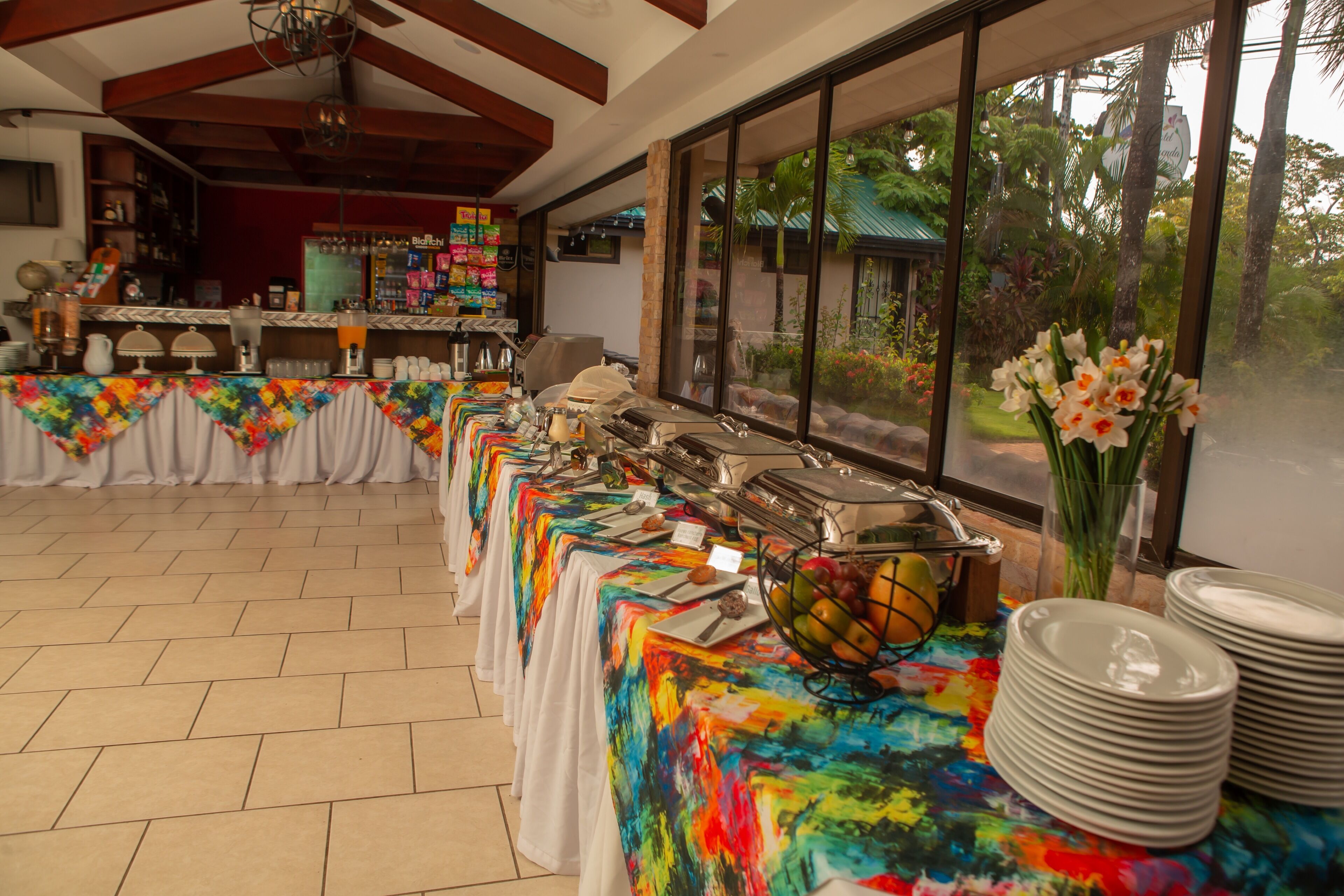 A buffet setup with a long table covered in a colorful tablecloth, a fruit bowl, and stacked plates.