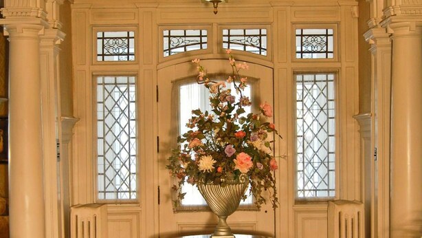 A grand hallway with a central round table topped with a floral arrangement, flanked by tall columns and large windows with intricate designs. - The Albanus Phillips Inn (Cambridge)