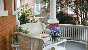 A wicker sofa with a cushion and a small table with a vase of flowers on a porch with a white railing. - The Albanus Phillips Inn (Cambridge)