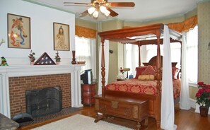A four-poster canopy bed with a red and gold patterned bedspread, a wooden bench at the foot of the bed, a brick fireplace with a screen, and a ceiling fan. - The Albanus Phillips Inn (Cambridge)