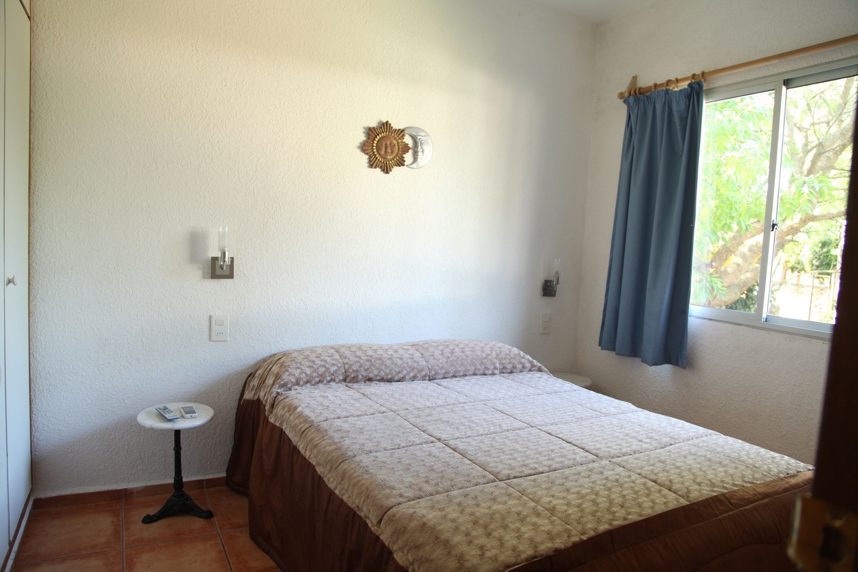 Quarto com cama, uma mesinha e janela com cortinas.
