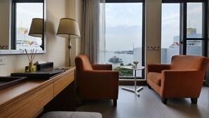 A hotel room with two orange armchairs, a wooden desk, a lamp, and a view of a body of water with a ship. - Watermark Hotel Sizihwan (Kaohsiung)