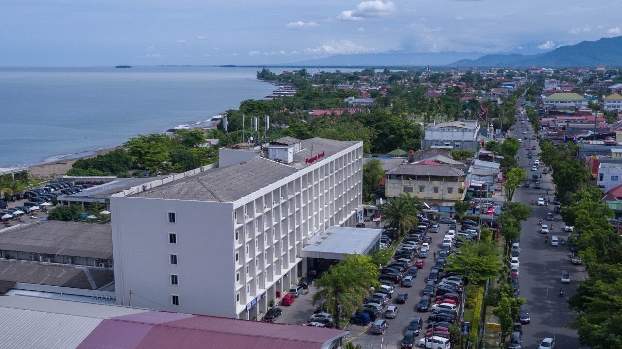 A coastal hotel with multiple floors, a parking lot, and a view of the sea and mountains in the background.