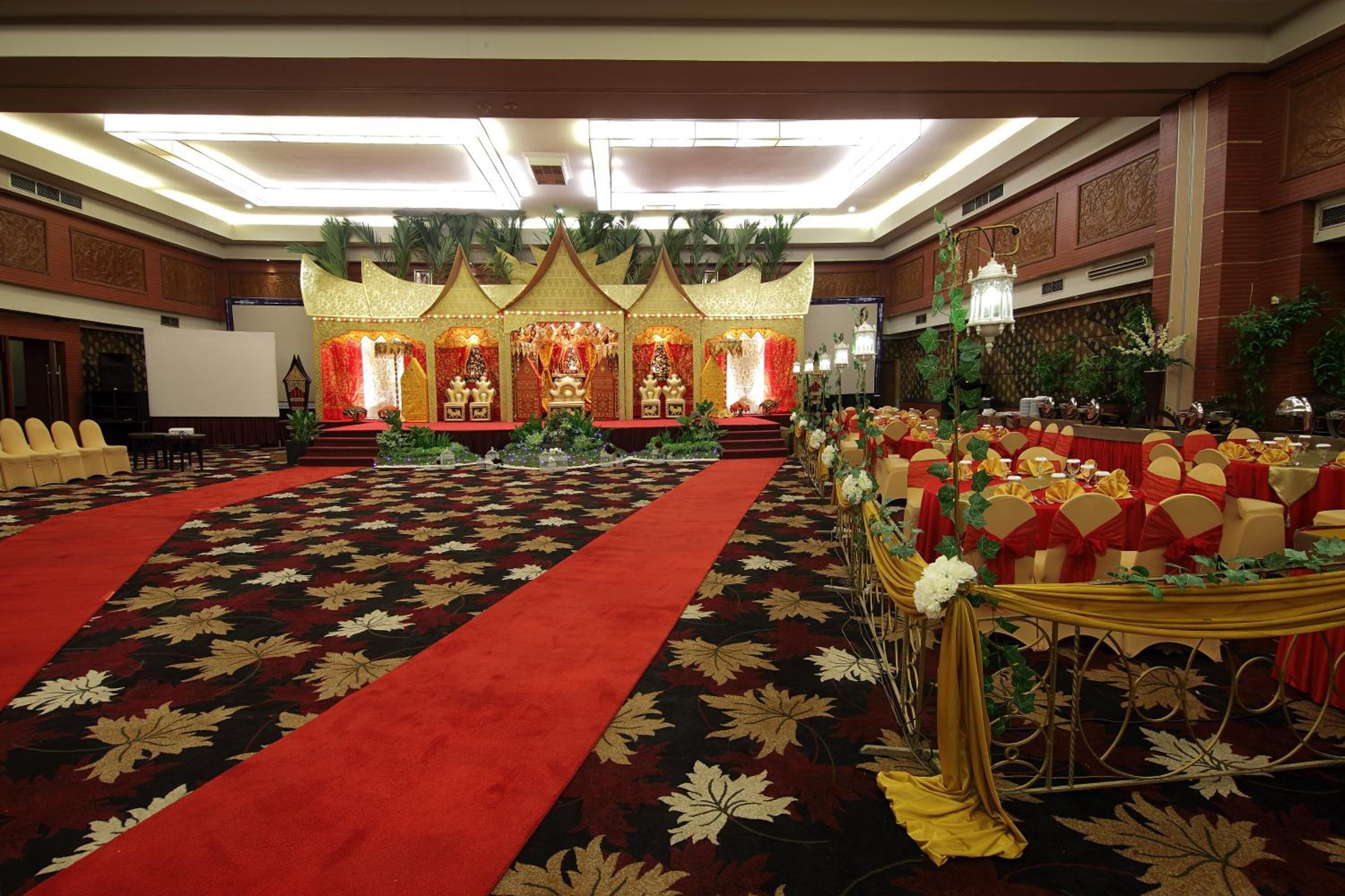 A banquet hall with a red carpet, tiered stage, and rows of tables with yellow chairs.
