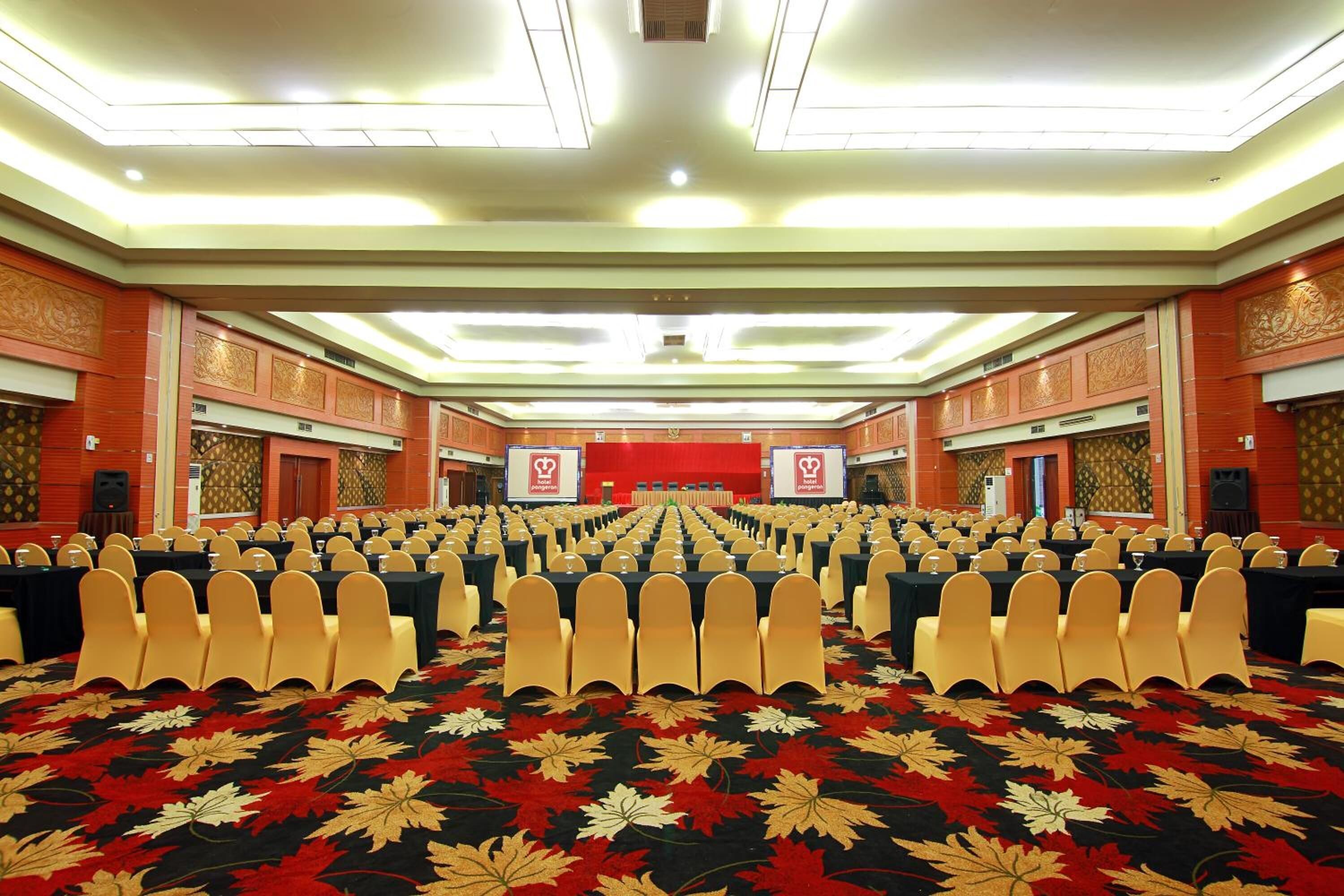 Un amplio salón de conferencias con filas de mesas y sillas, una alfombra roja y un escenario al frente.