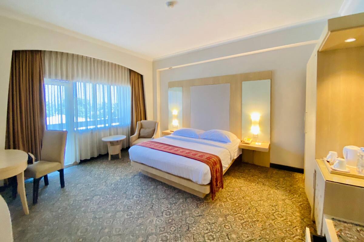 A hotel room with a bed, a chair, a small table, a desk with a telephone, and a window with curtains.