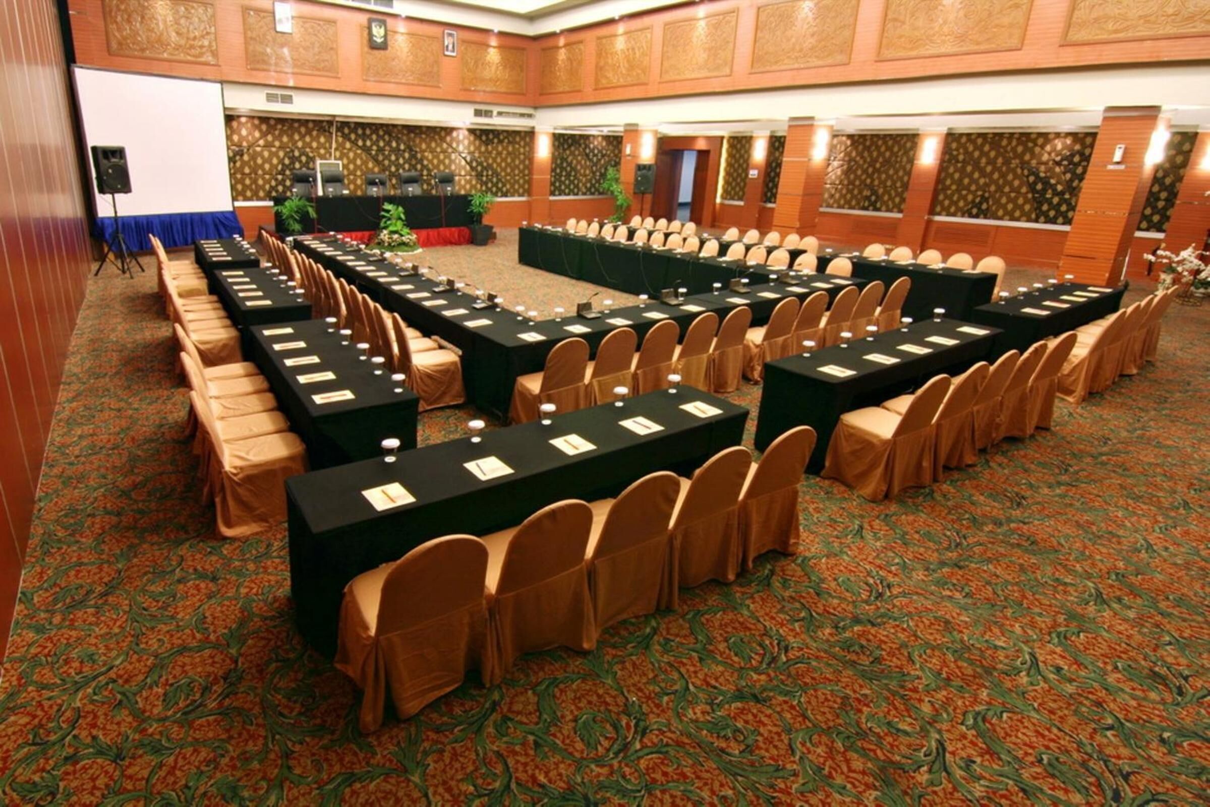 A conference room with long tables and chairs, a stage with a podium, and a carpeted floor.
