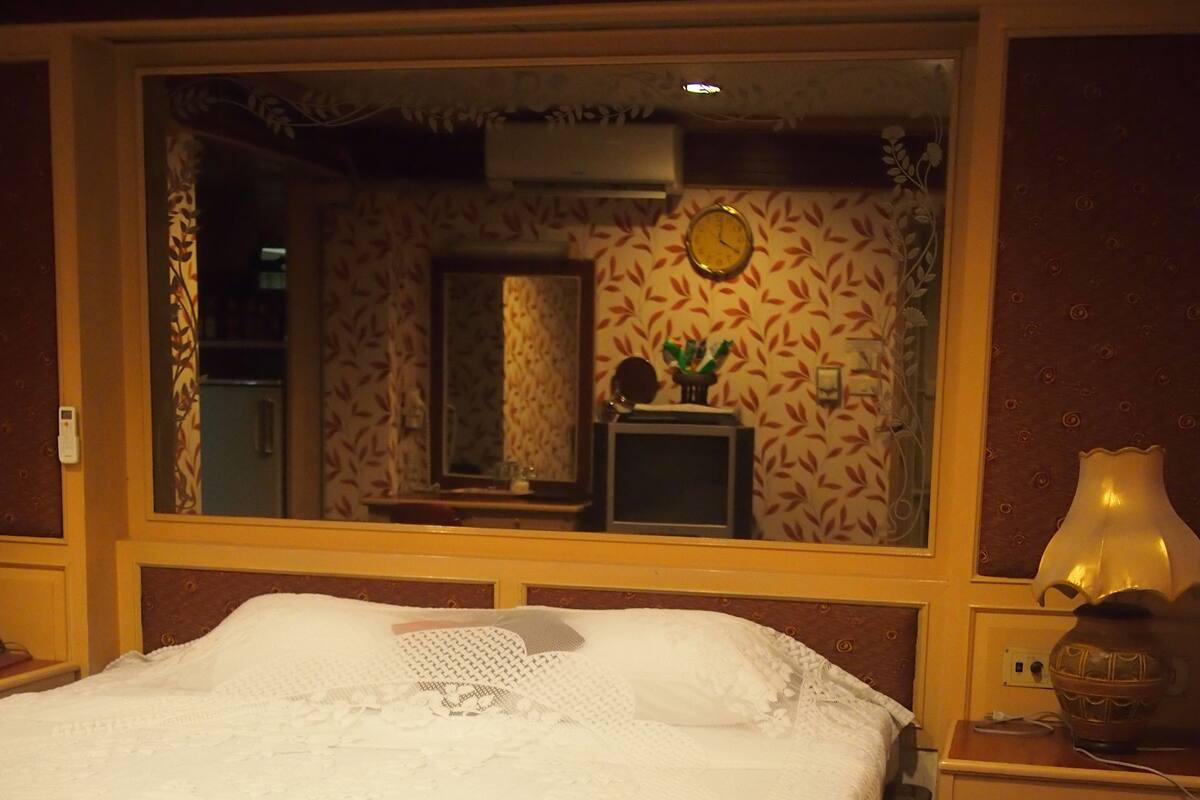 A bed with a white bedspread and a decorative headboard. There is a nightstand with a lamp and a vase. A mirror is on the wall above the bed.