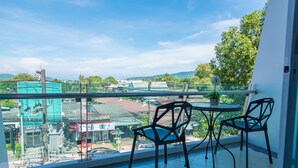 A balcony with two chairs and a table, overlooking a cityscape with buildings and greenery. - Phuket Jula Place (Chalong)