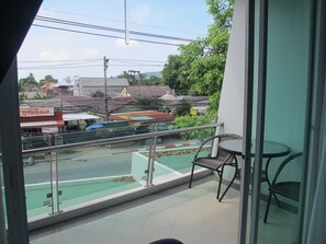 A balcony with a view of a street and buildings, a small table, and a chair. - Phuket Jula Place (Chalong)