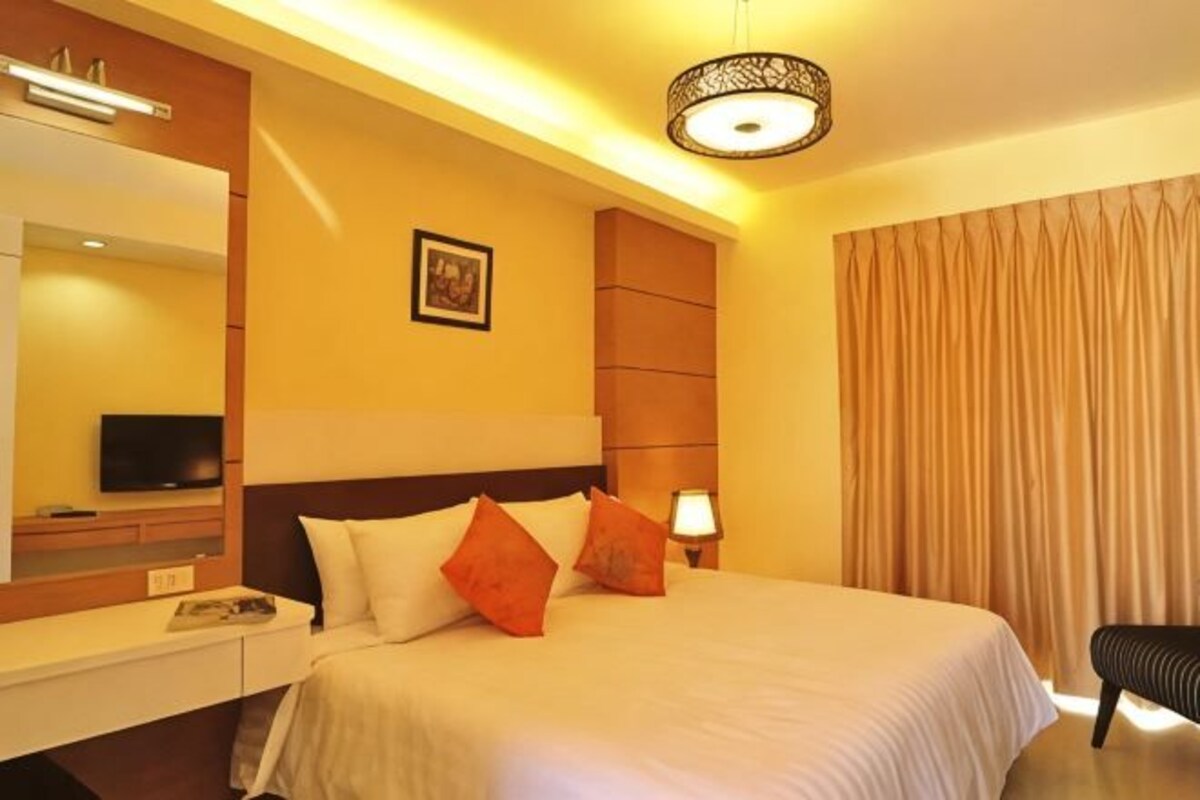 Una habitación de hotel con cama, mesita de noche, lámpara, televisor y un gran ventanal con cortinas.