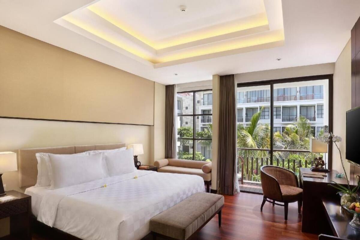 Ein Hotelzimmer mit einem großen Bett, einem Schreibtisch, einem Stuhl und einem Balkon mit Blick auf Gebäude und Grünflächen.
