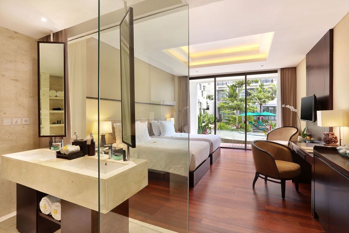A hotel room with a large bed, a desk, a chair, a bathroom with a mirror, and a view of a pool and buildings.