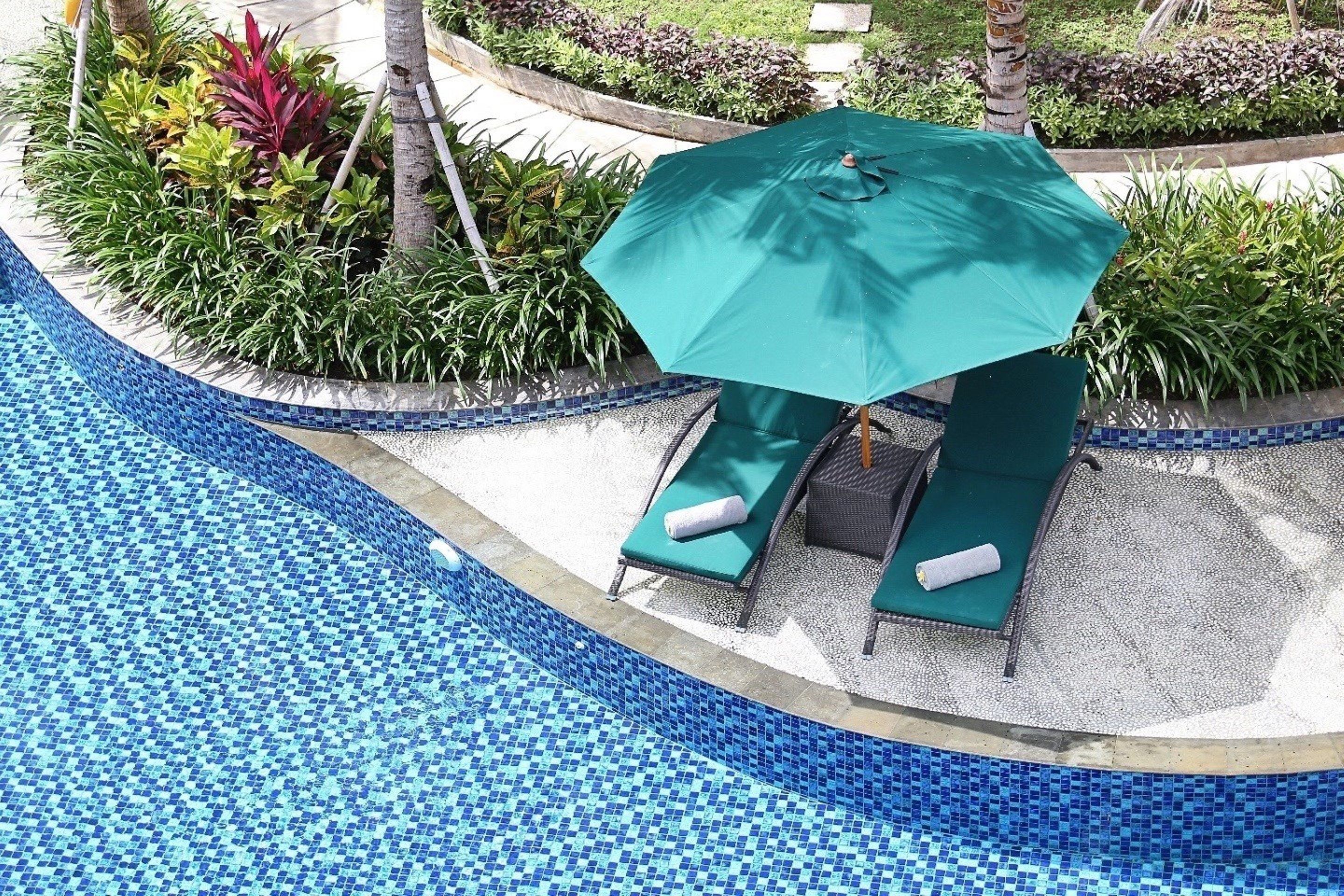 A poolside area with a green umbrella, two lounge chairs, and a small garden with various plants.