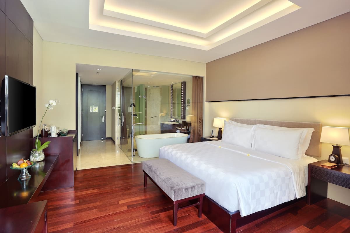 Une chambre d’hôtel moderne dotée d’un grand lit, d’une télévision, d’un bureau agrémenté d’un vase de fleurs et d’une salle de bain visible à travers une porte en verre.
