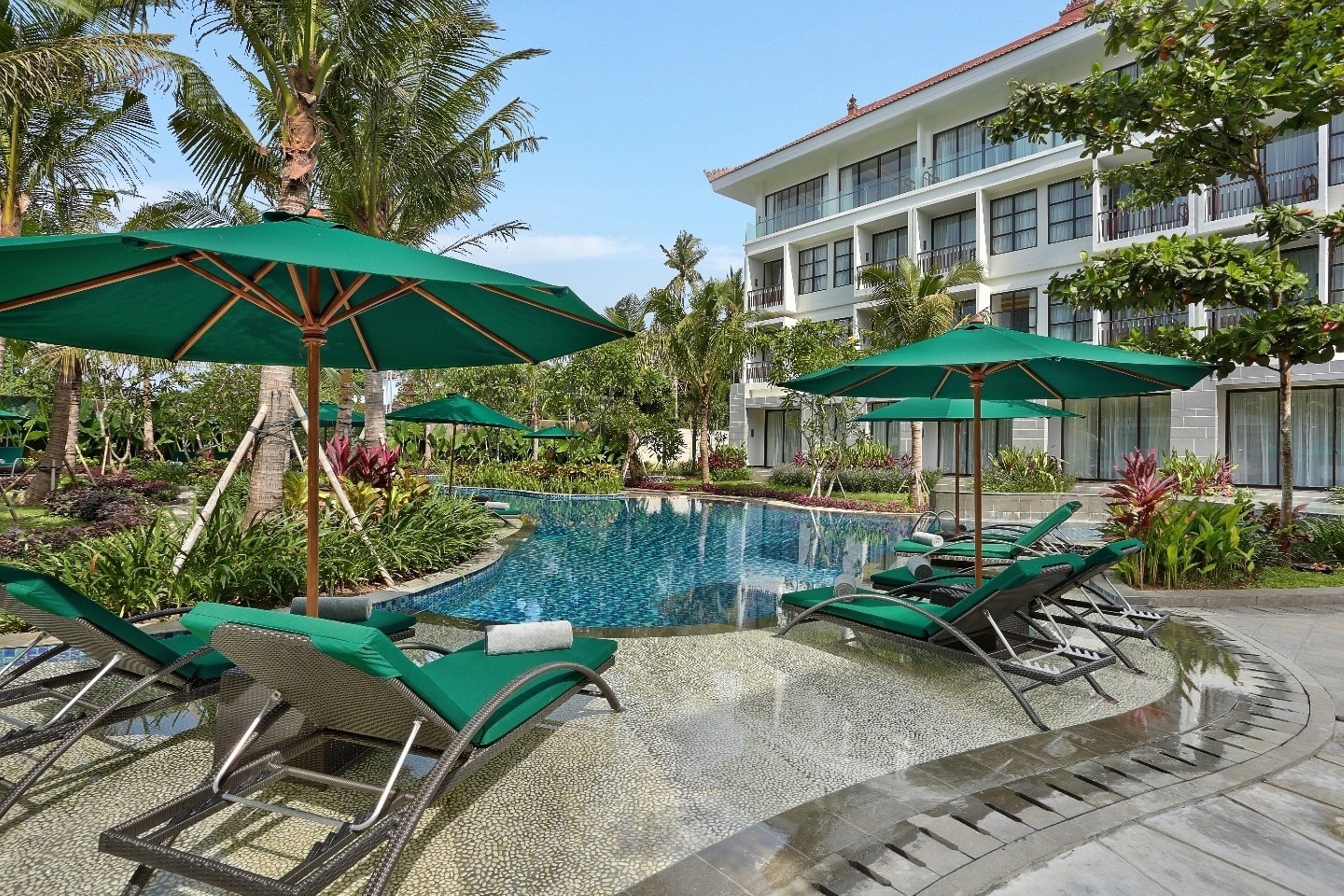 A pool area with lounge chairs and umbrellas, surrounded by palm trees and a building with balconies.