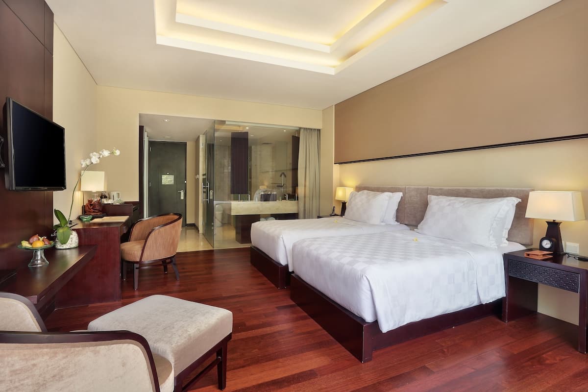 Une chambre d’hôtel moderne dotée d’un grand lit, d’un bureau avec un téléviseur à écran plat, d’un coin salon avec des fauteuils et d’une salle de bain visible à travers une porte en verre.