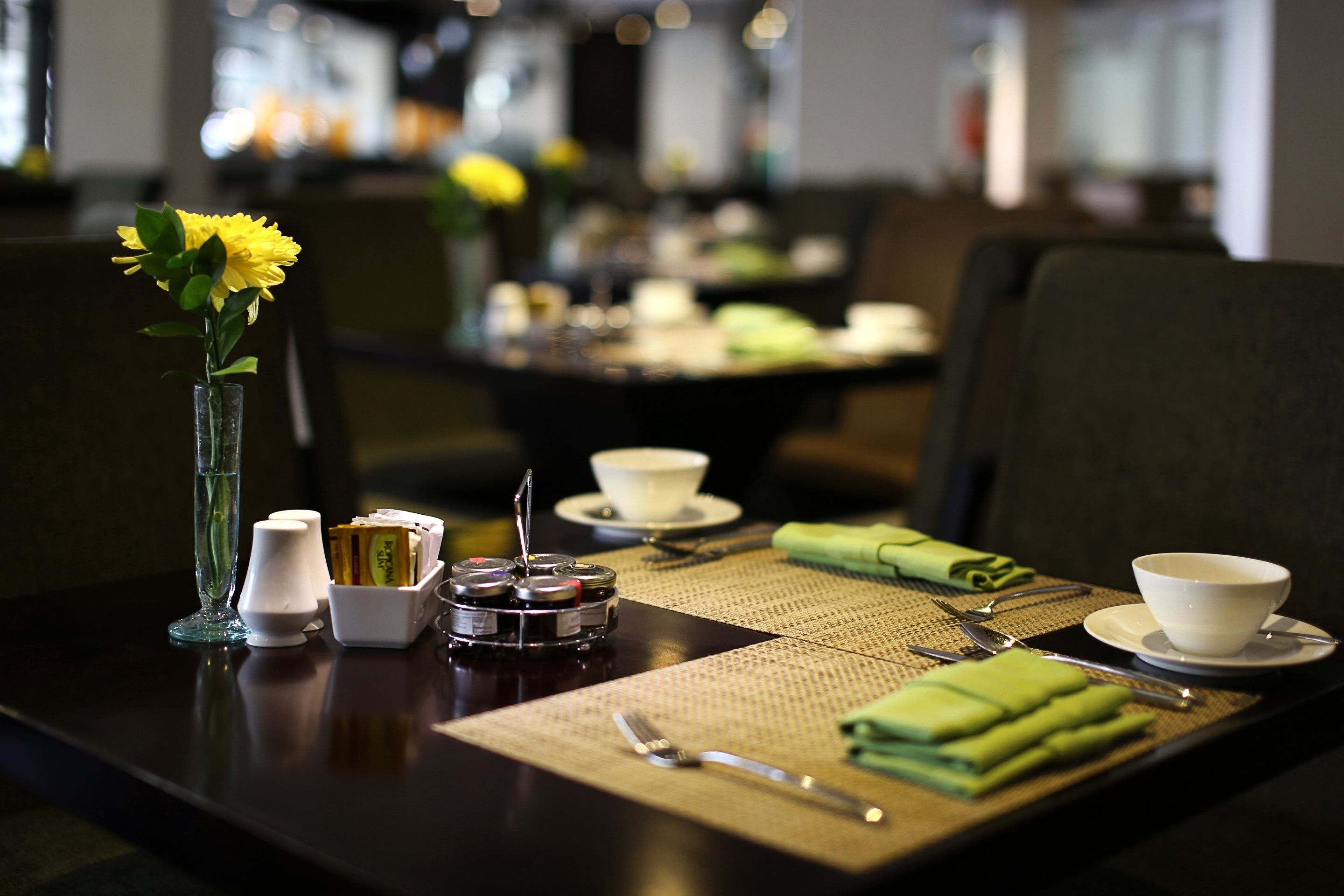 A dining table set with a vase of yellow flowers, condiments, and cutlery.