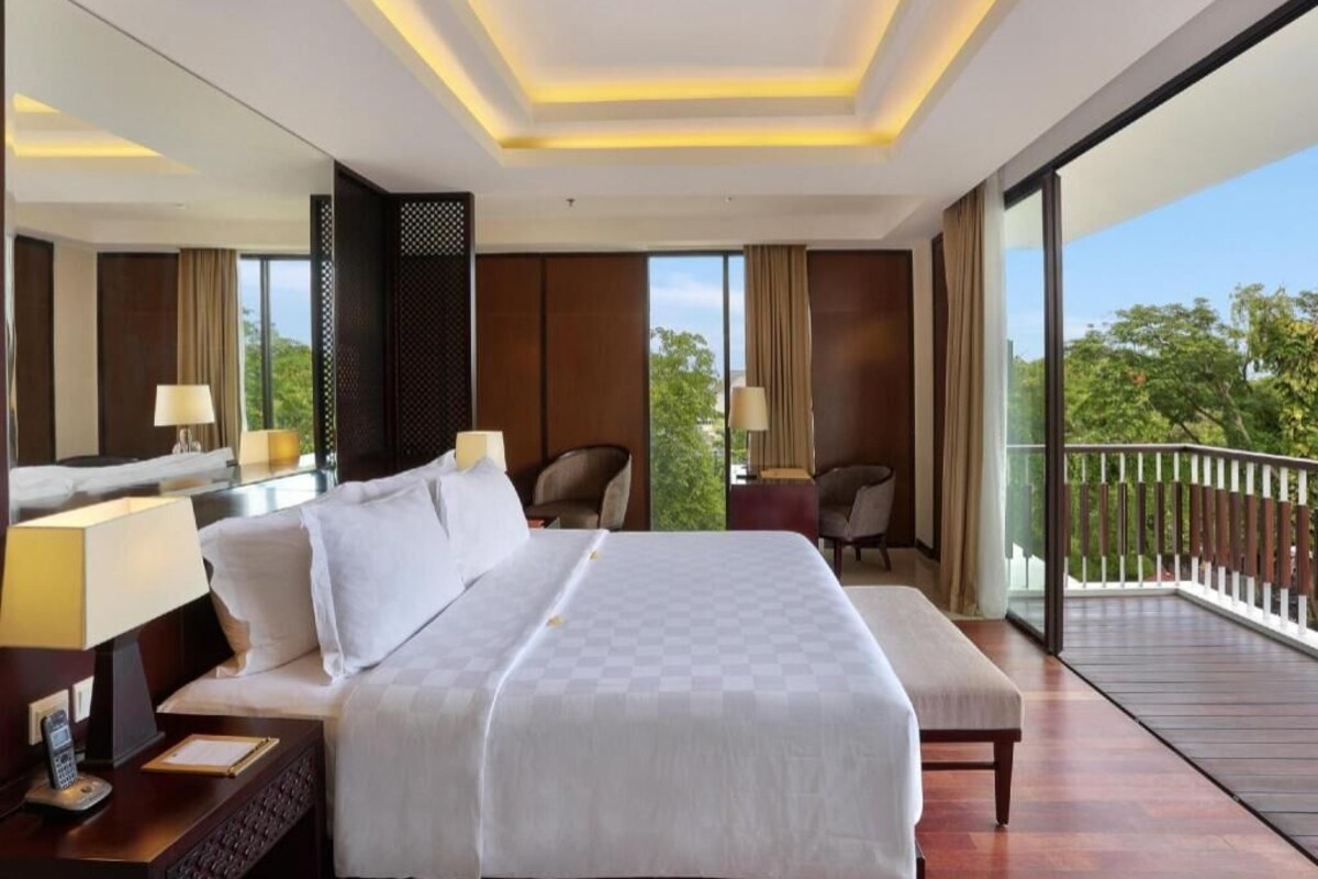A hotel room with a large bed, bedside tables, a desk, a chair, a balcony with a railing, and a view of greenery.
