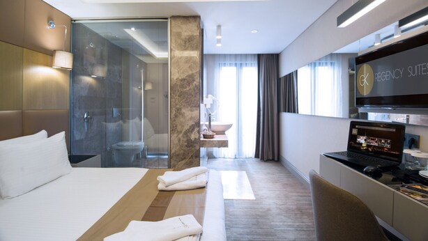 A modern hotel room with a large bed, a desk with a laptop, a glass shower enclosure, and a view of the city. - GK Regency Suites (Istanbul)