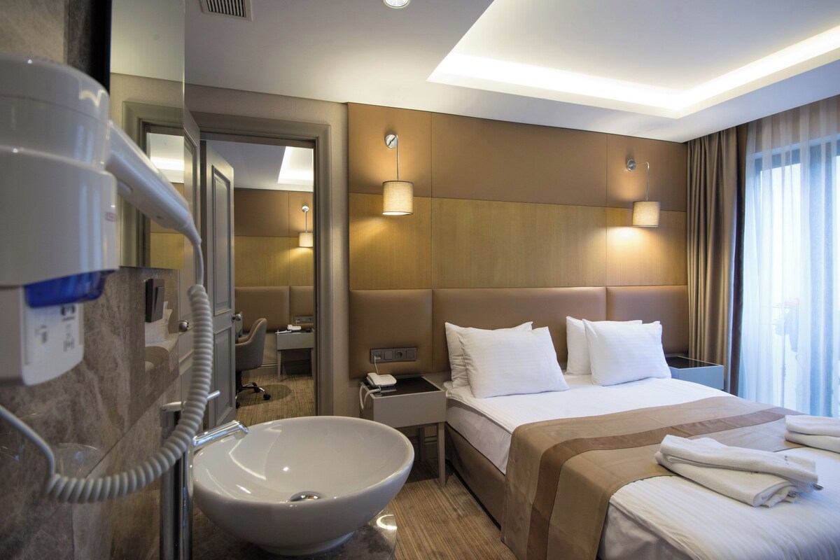 A hotel room with a large bed, bedside tables, a bathroom with a sink and mirror, and a view of the city through the window.