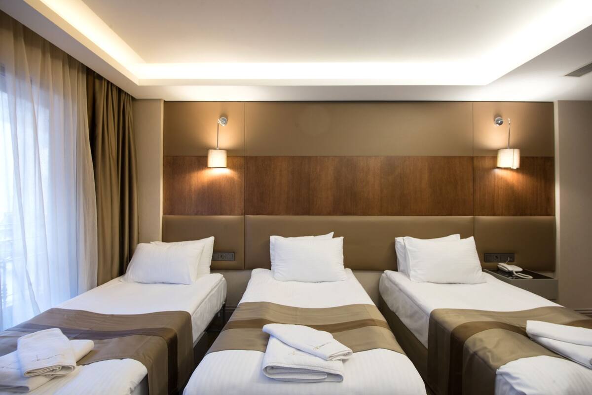 A hotel room with three single beds, each with a brown headboard and white bedding. There are two wall-mounted lamps on either side of the beds.