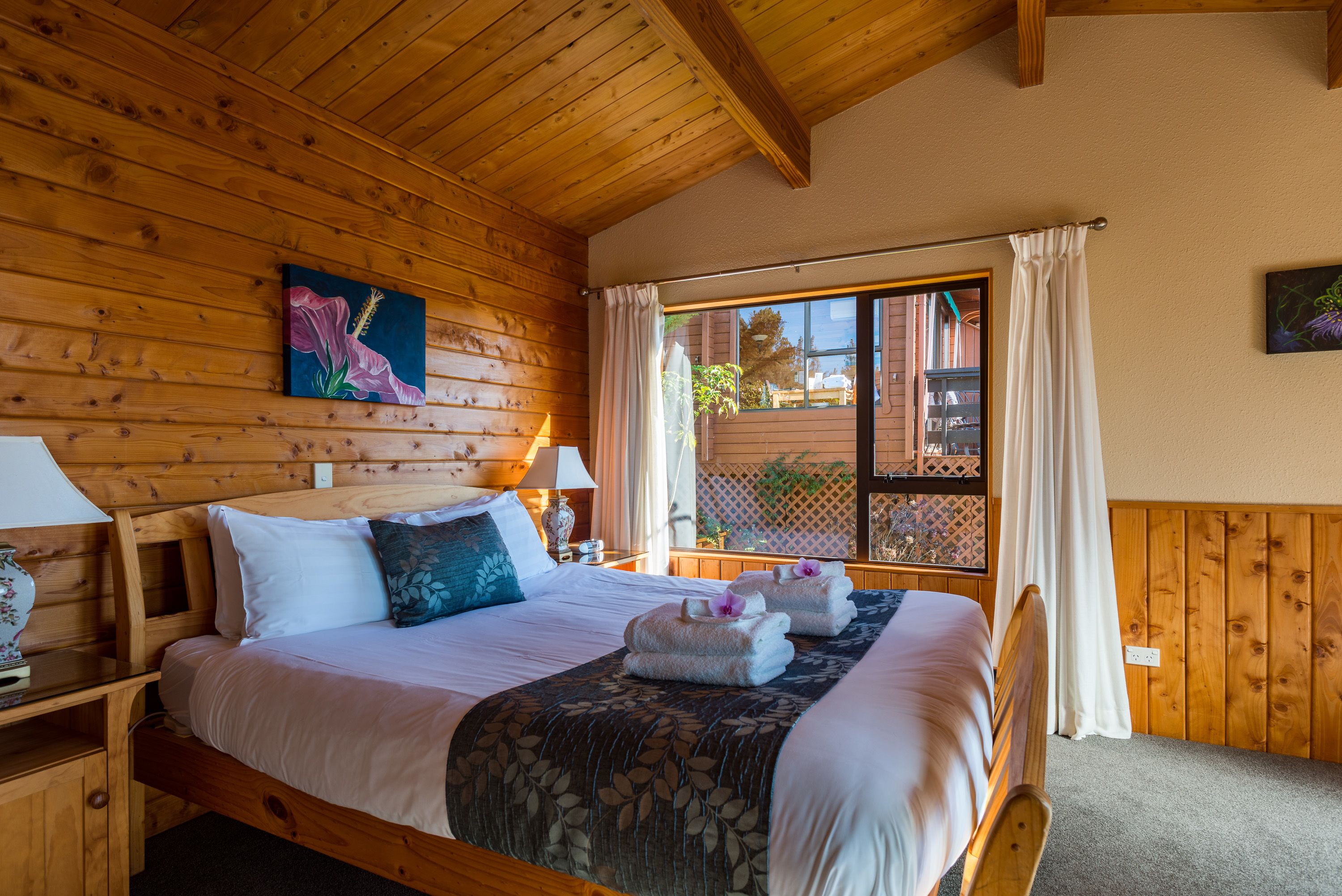 A wooden cabin-style bedroom with a large bed, two bedside tables, a window with curtains, and a painting on the wall.