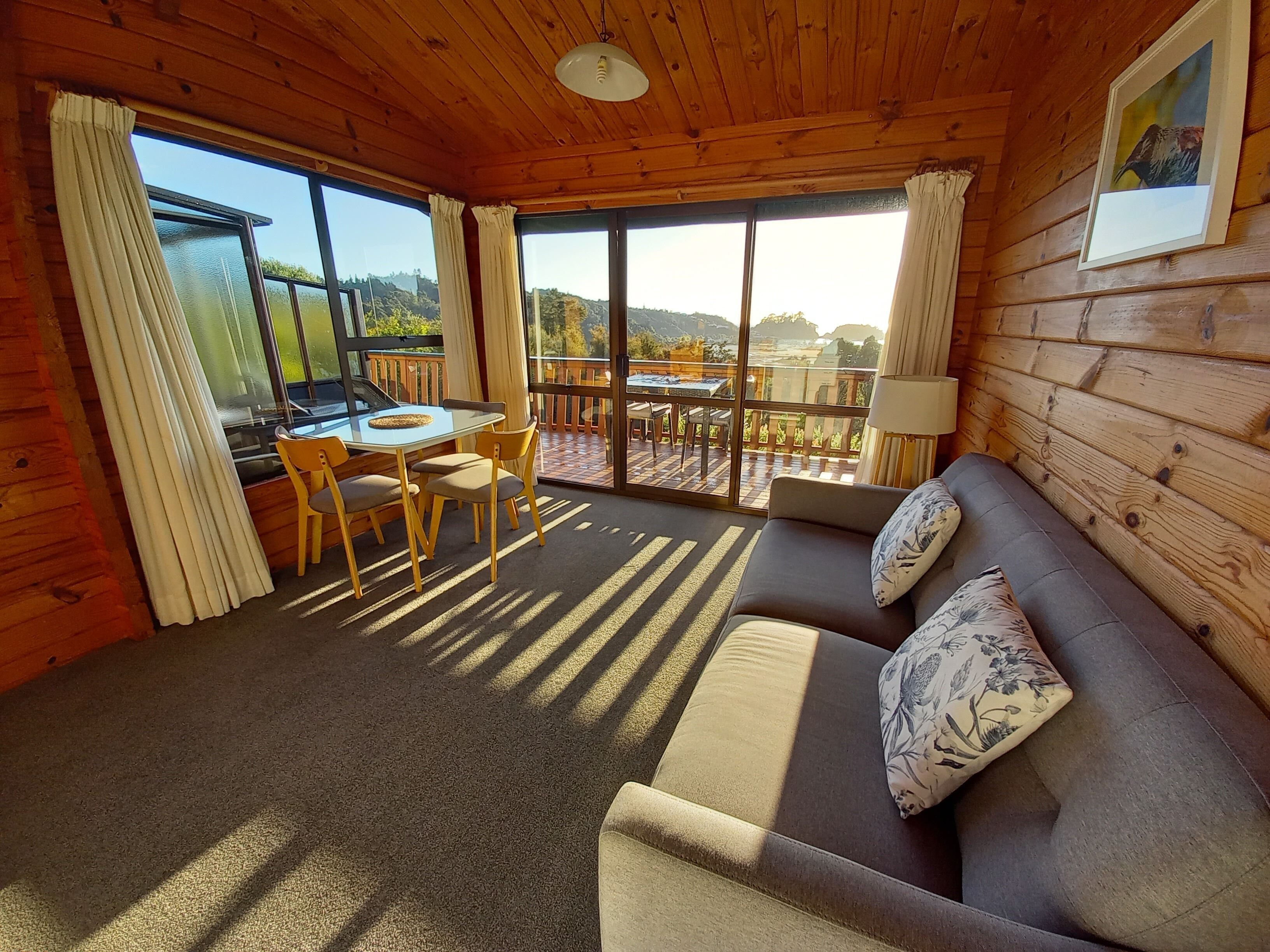 A cozy cabin interior with a wooden ceiling, a sofa with floral cushions, a dining table with chairs, and large windows offering a view of the outdoors.