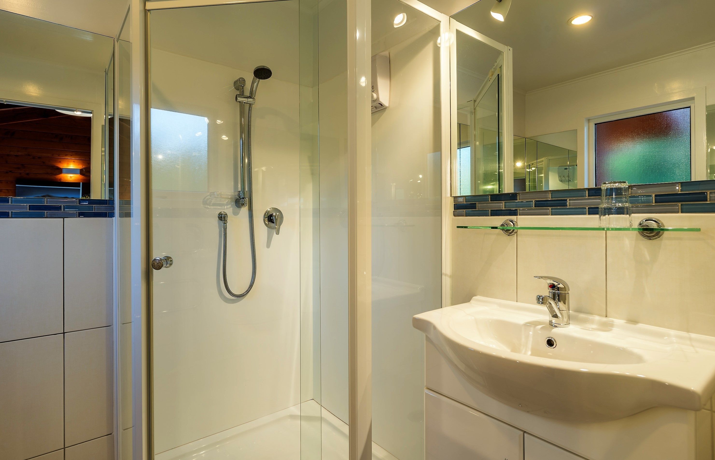 Une salle de bain moderne avec une douche à parois de verre, un lavabo blanc et un miroir agrémenté d’un pare-splash en carreaux bleus.