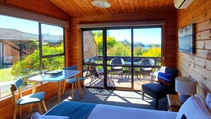 A wooden cabin room with a bed, a table, and chairs. There is a large window with a view of greenery and a building. - Kimi Ora Eco Resort (Kaiteriteri)