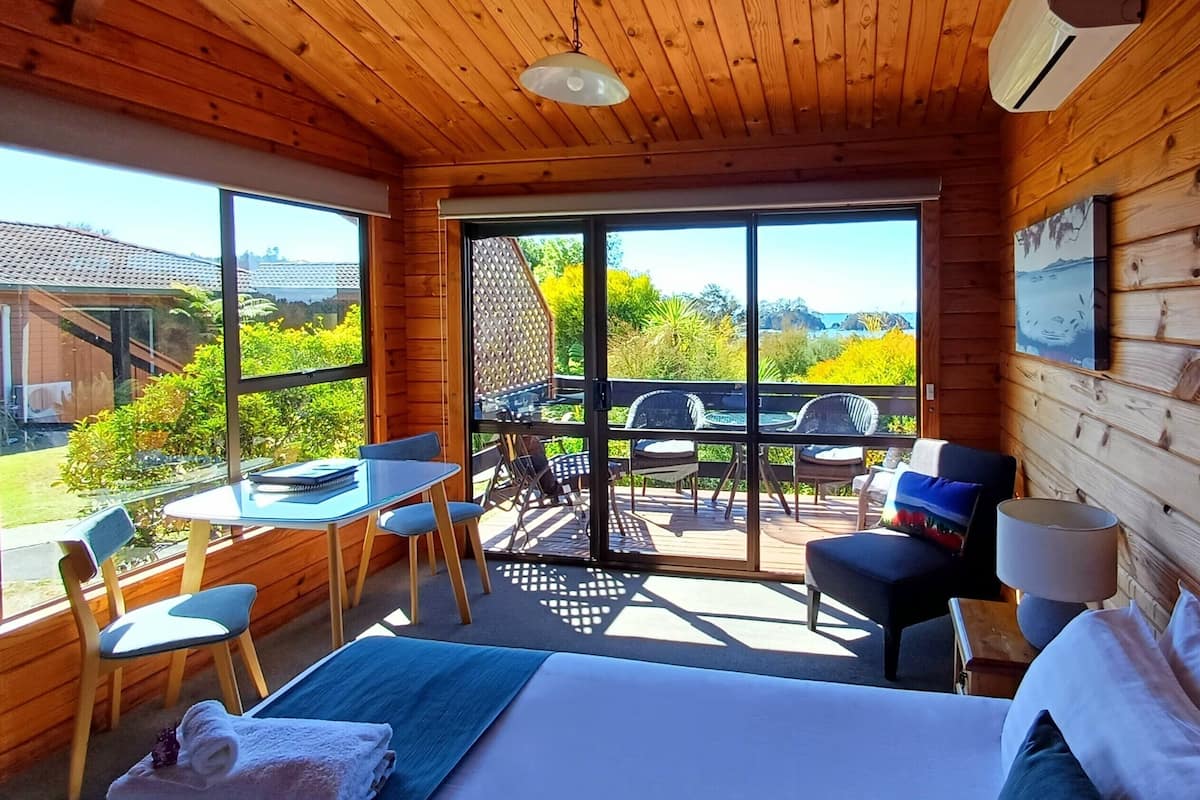 A wooden cabin room with a bed, a table, and chairs. There is a large window with a view of greenery and a building.