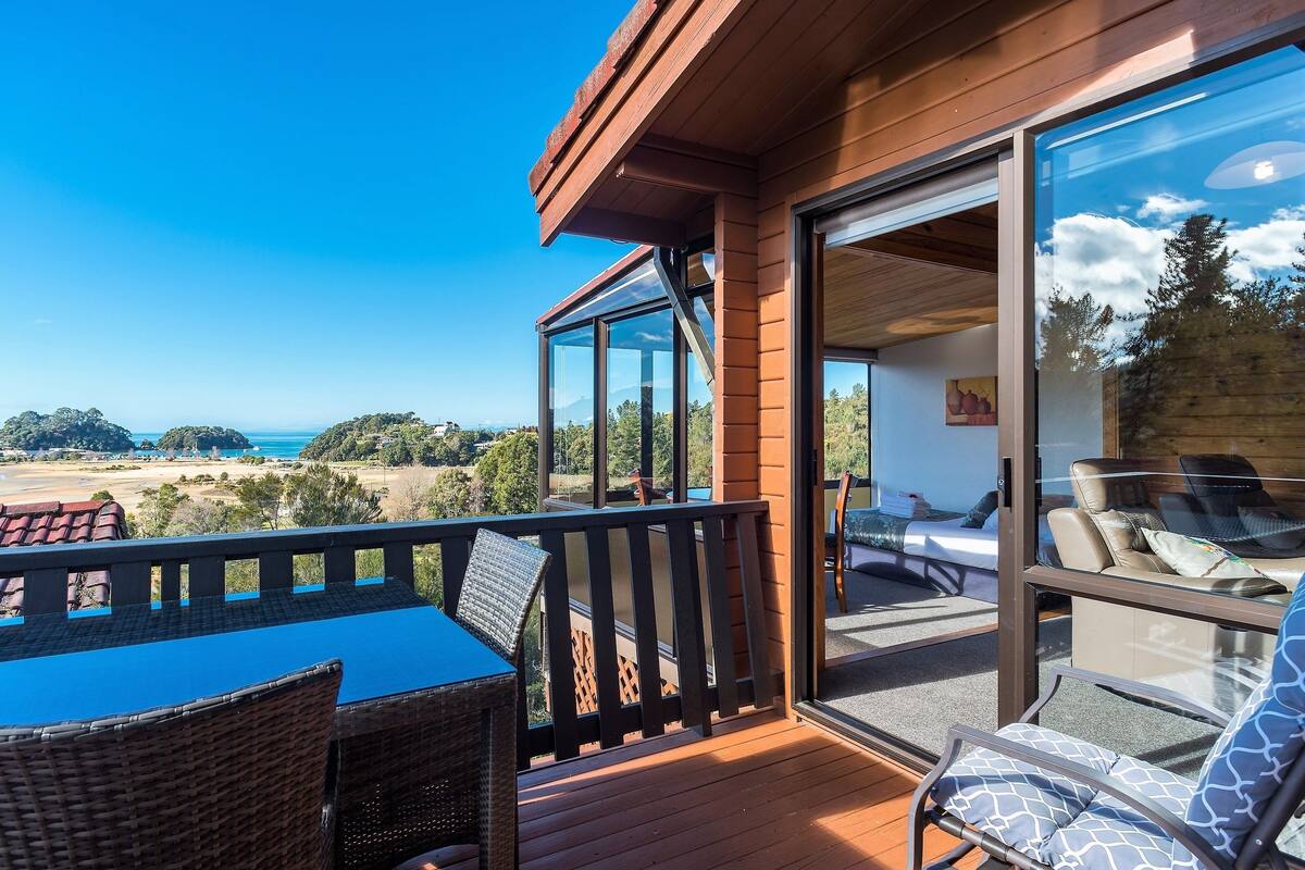 A balcony with wicker furniture, a glass door leading to a room with a bed and a painting, and a view of the ocean and hills.