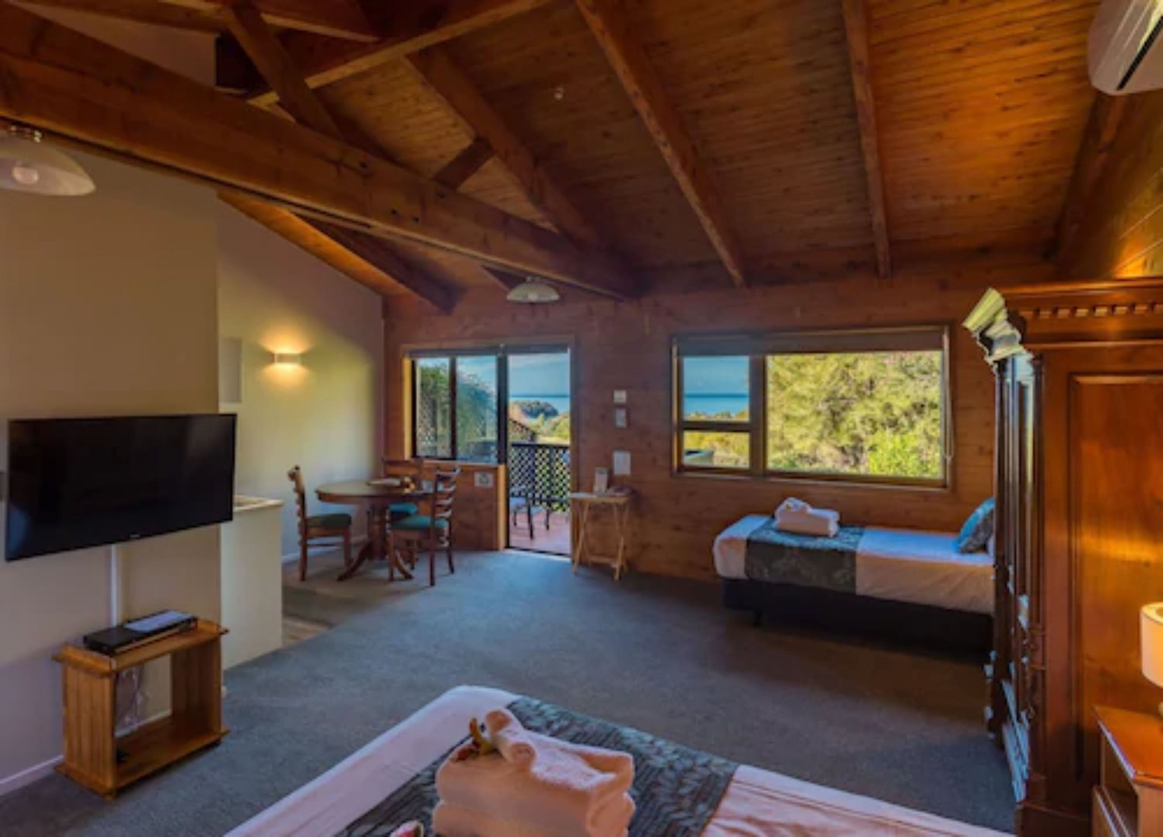 A wooden cabin interior with a bed, a TV, a dining area, and a balcony with a view.