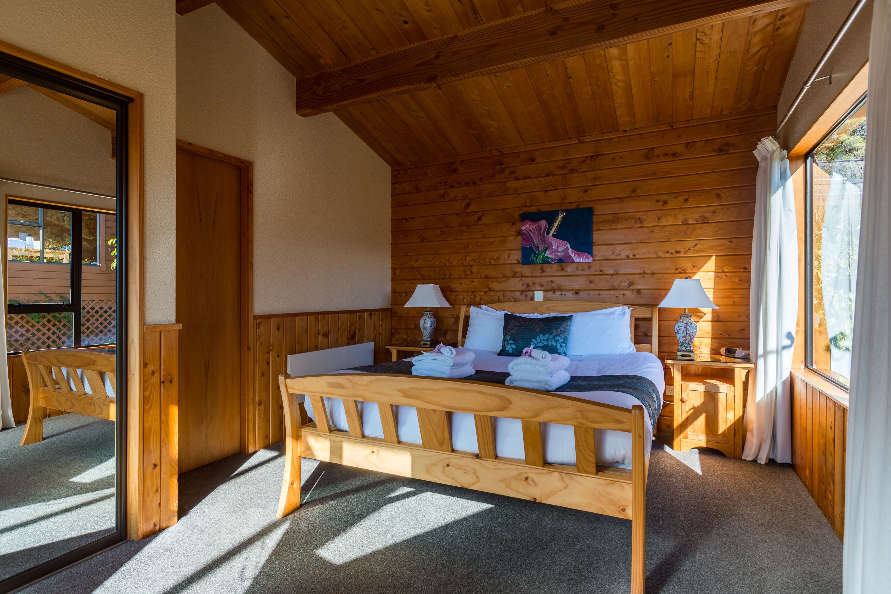 A wooden-framed bedroom with a bed, two bedside lamps, a painting on the wall, and a window with curtains.