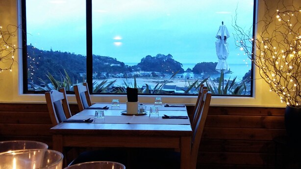 A dining table with chairs and candles, a window with a view of a beach and ocean, and decorative lights. - Kimi Ora Eco Resort (Kaiteriteri)