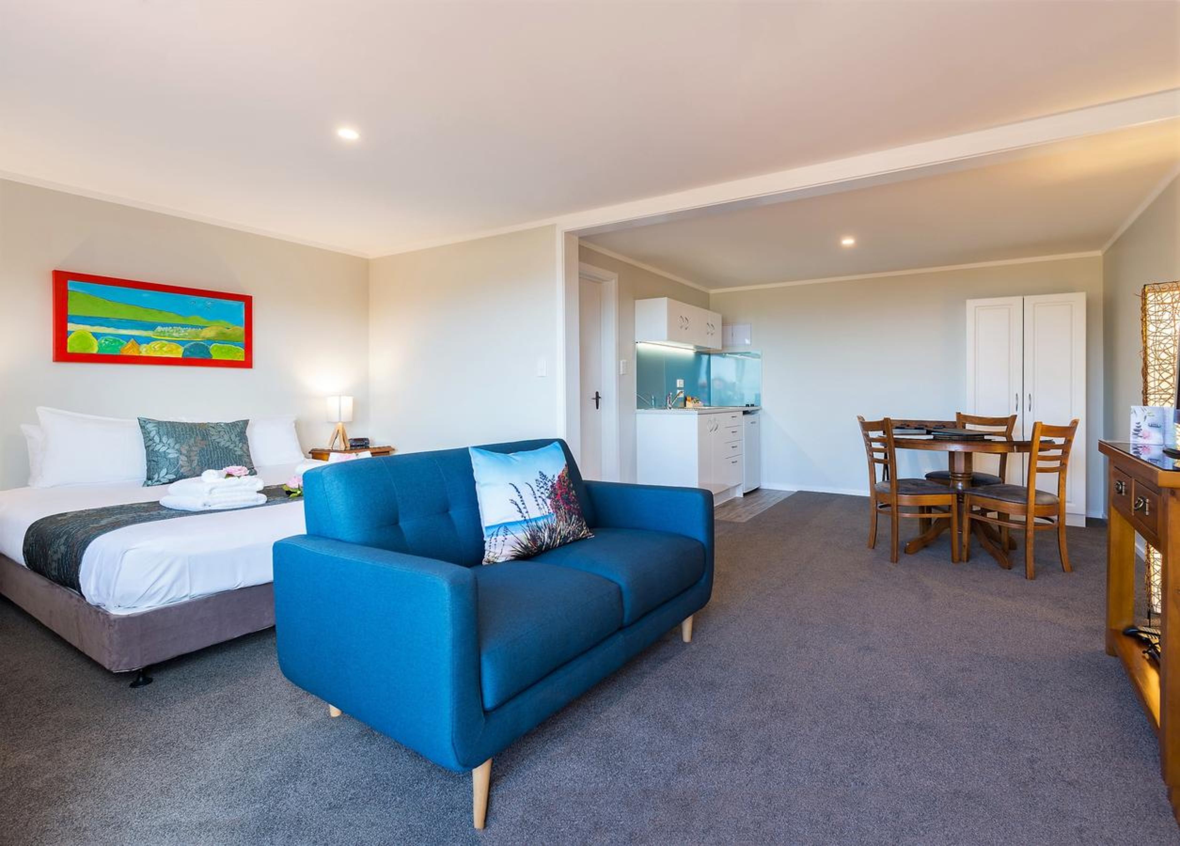 Una habitación de hotel con cama, sofá azul, mesa de comedor con sillas, kitchenette y un cuadro en la pared.