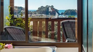 A bathroom with a bathtub, a window, and a view of a balcony with chairs and a railing. - Kimi Ora Eco Resort (Kaiteriteri)