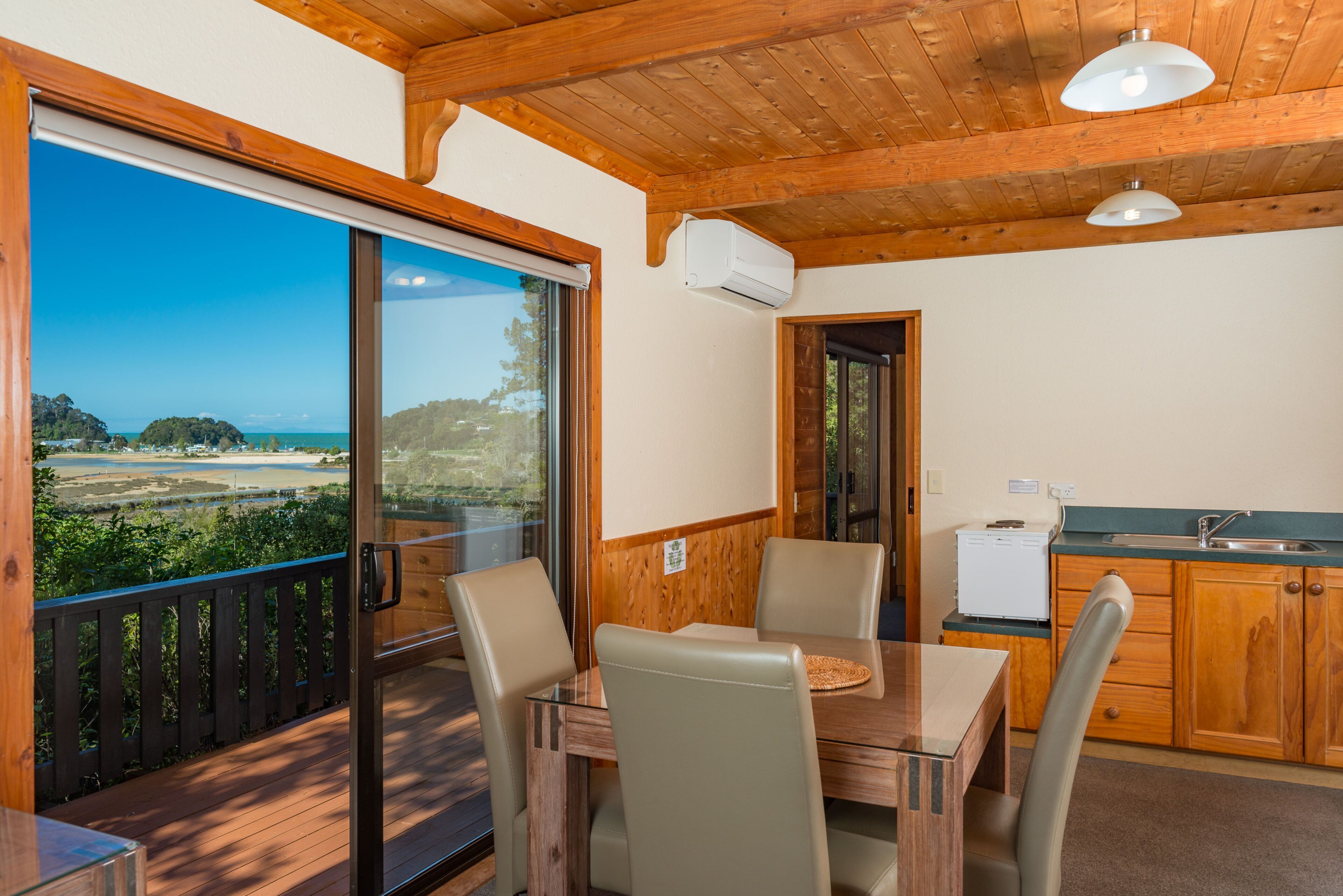Una terraza de madera con una mesa y sillas, una cocina con armarios de madera, y vistas a un paisaje costero a través de amplios ventanales.
