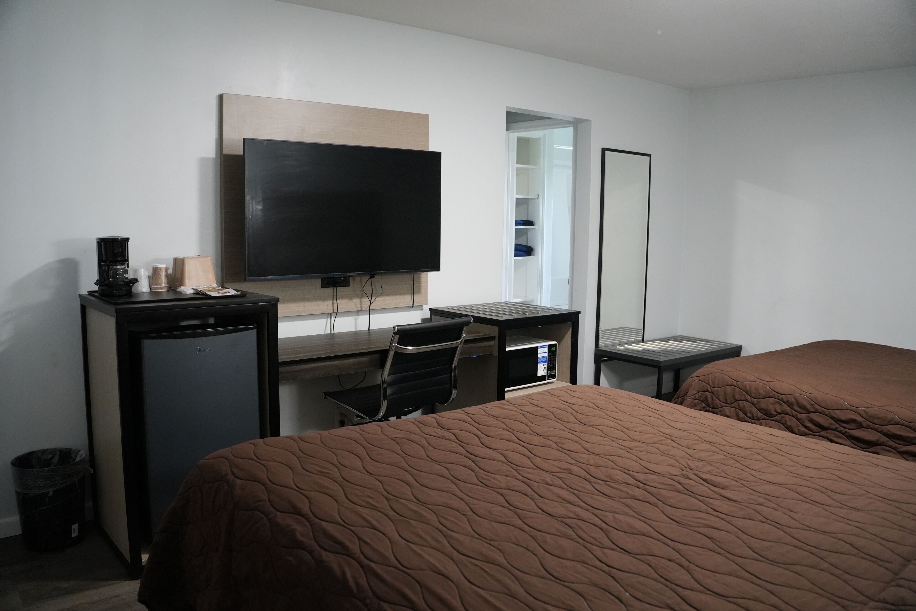 A hotel room with a bed, a desk with a computer, a flat-screen TV, a mirror, and a small refrigerator.